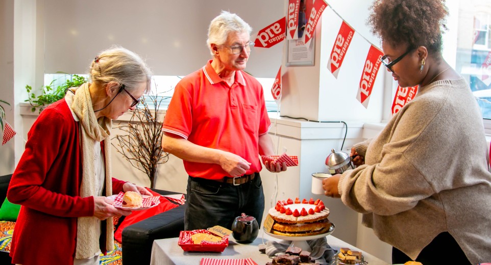 Two women and a man stood together. One woman is pouring tea for the man, whilst the other is putting cake on her plate. They are stood around a table full of baked goods.