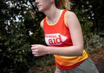 A woman running. There are trees behind her, and she's wearing and red and yellow Christian Aid running vest.