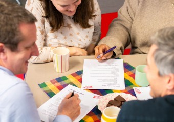 A table displaying brownies and cups of tea. A group of people are leant on the table, filling in a Christian Aid quiz.